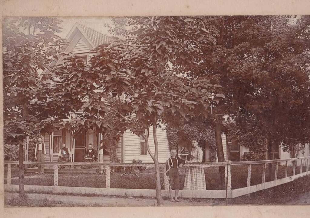 People sitting on house front porch and standing in front yard and trees with full leaf canopies