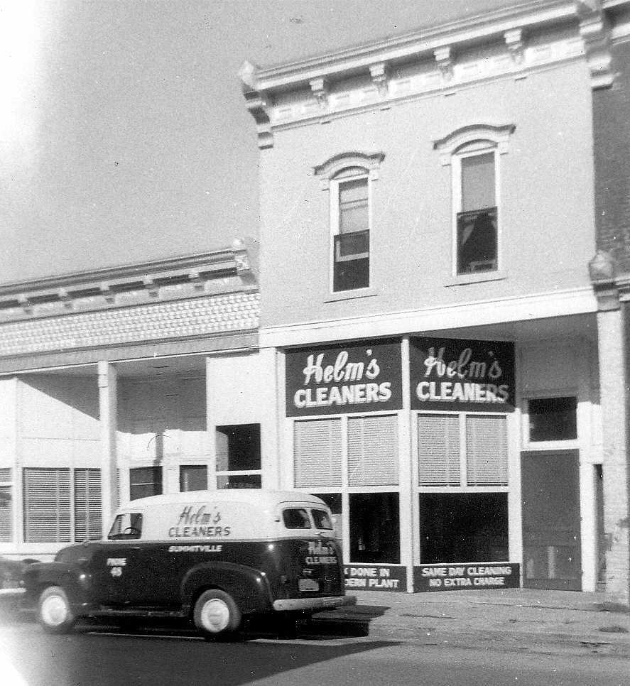 Helm's Cleaners storefront with a car parked in front of it with a white upper half and a black lower half and the words "Helm's Cleaners" painted on it