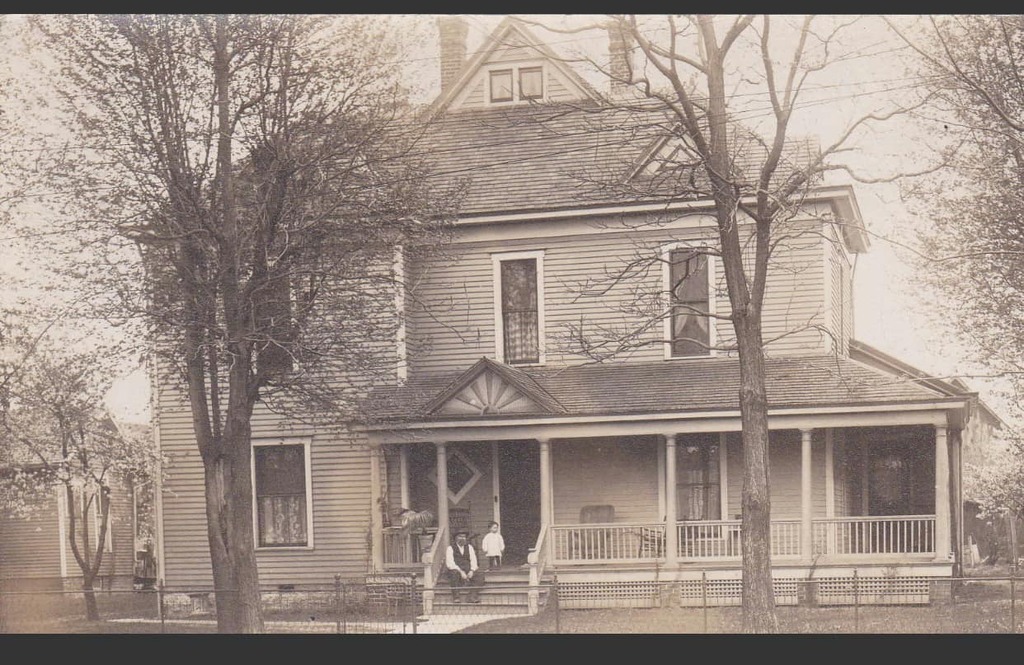 Man and child on front porch of two story house and two large trees in yard